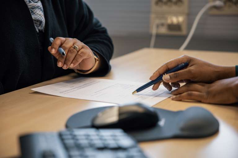 Kaksi henkilöä istuu pöydän ääressä, molemmat pitelevät kädessään kynää ja tarkastelevat samaa paperia. Pöydän etualalla näkyy tietokoneen hiiri ja näppäimistö.