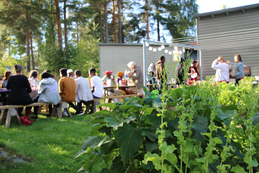 Etualalla on viljelylaatikossa kasvavia vihanneksia, taustalla joukko ihmisiä istuu pitkän puutarhapöydän ääressä.