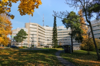 A light-colored multi-story building with many windows. The building is surrounded by trees in yellow and orange autumn colors. In the foreground, there are parking spaces, a lawn area, a walking path, and a bicycle shelter.