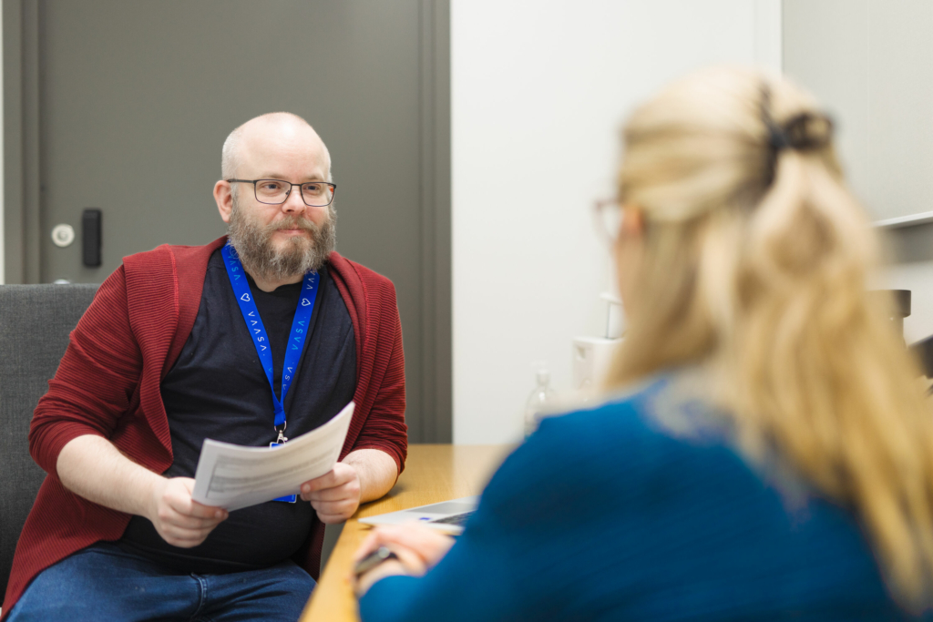 A customer service representative and a customer sit at a table in an office space. The representative is holding papers and listening to the customer.