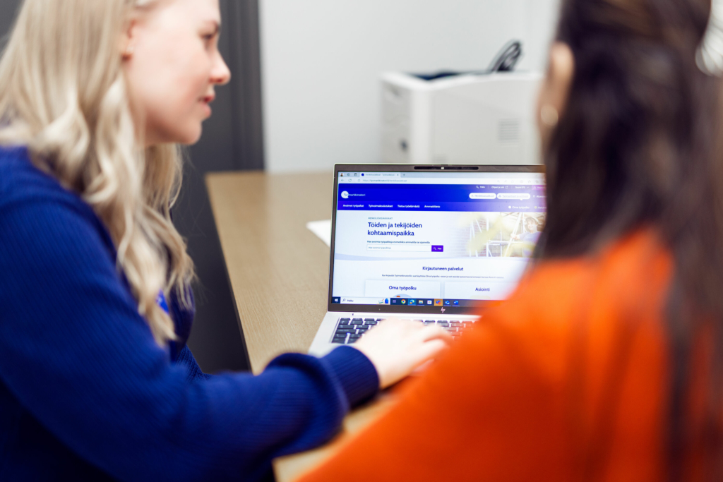 Two people are using a laptop at a table. The person on the left is typing something on the keyboard, and the screen displays the Job Market Finland website.