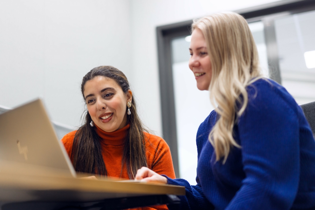 Two young people sit at a laptop, smiling as they look at the screen.