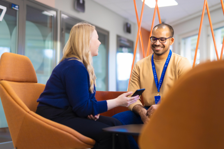 Two people sit in a bright lobby area in armchairs. The customer shows something on their phone to the customer service representative, and they smile.