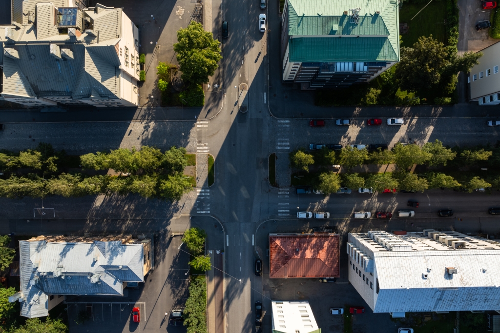 An aerial view of a street scene showing an intersection, pedestrian crossings, cars parked and in motion, and lush trees lining the streets. The buildings cast shadows on the street, and the photo is taken when the sun is low.