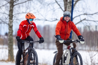 Two smiling cyclists are riding side by side in a snowy park landscape.