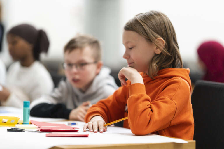 Close-up of a smiling 5th grade boy sitting at a table. In front of him there are some craft supplies. Behind him, the blurred silhouettes of two other children can be seen sitting at the table.