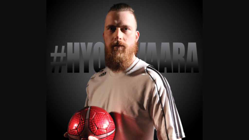 Tummalla taustalla teksti #hyokyvaara, etualalla mies jalkapalloasussa ja punainen jalkapallo kädessään.