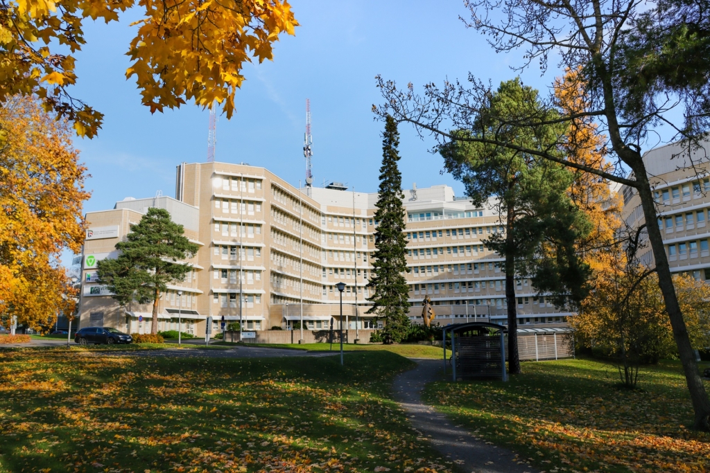 En ljus fler våningsbyggnad med många fönster. Byggnaden omges av träd i höstens gula och orangea färger. I förgrunden syns parkeringsplatser, ett grönområde, en gångstig och ett cykelskjul.